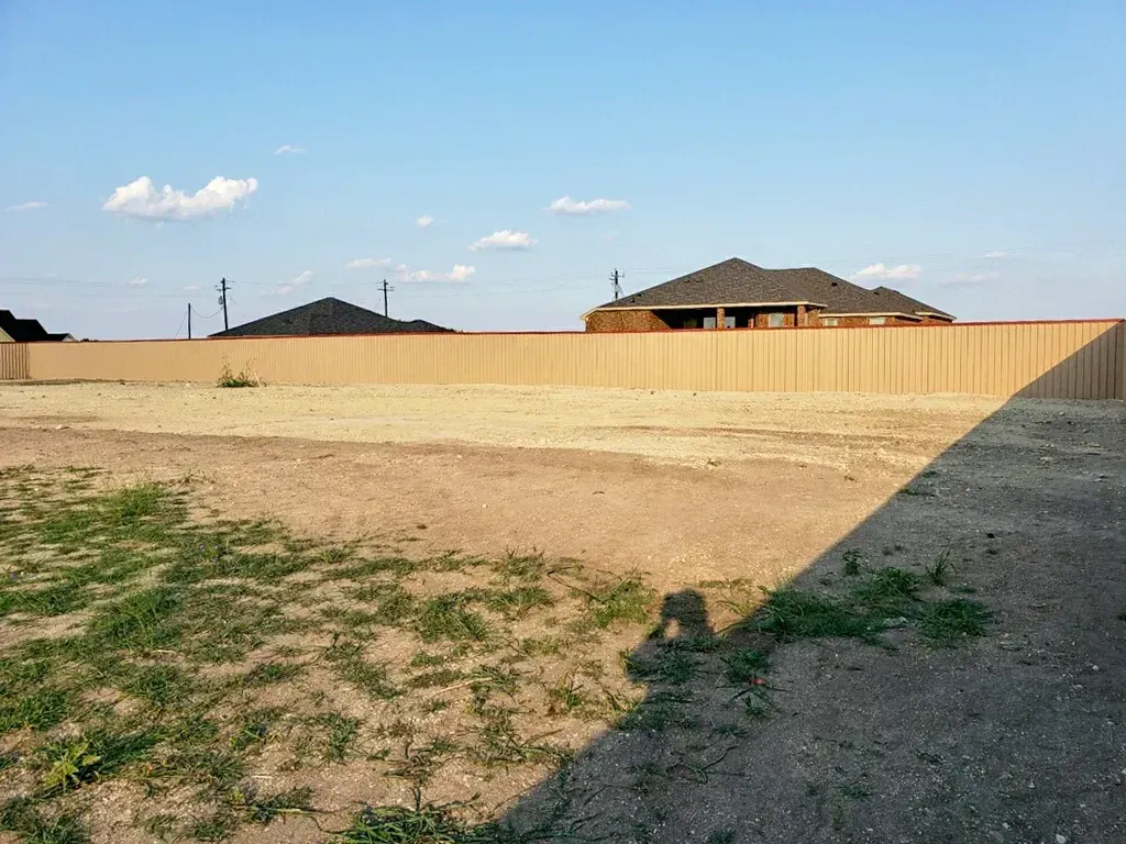 Empty lot with brown dirt and sparse grass, fenced in with a tan fence. Distant houses and blue sky.