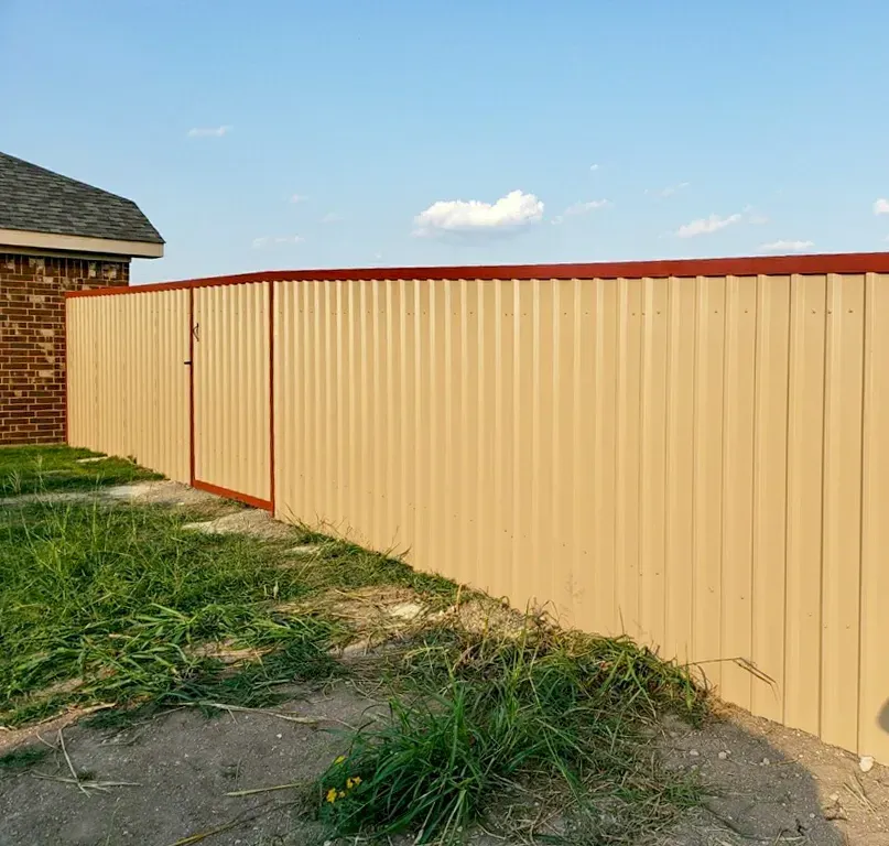 Beige corrugated metal fence with a red top, a gate, and green grass against a blue sky.