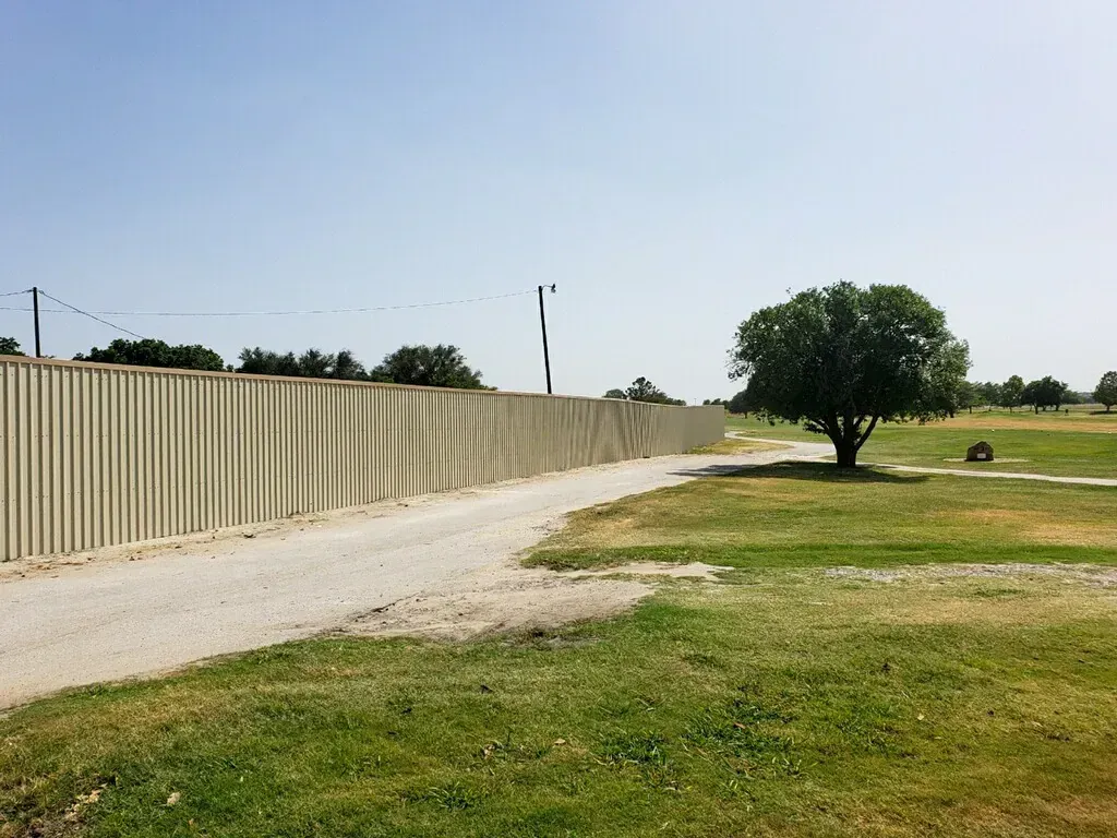 Gravel path alongside a tall, corrugated fence in a grassy park setting under a clear sky.