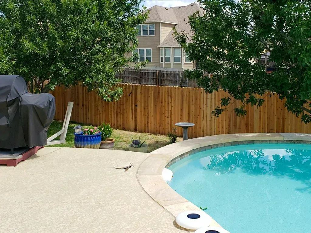 Backyard with a pool, fence, grill, trees, and a house in the background. Sunny day.