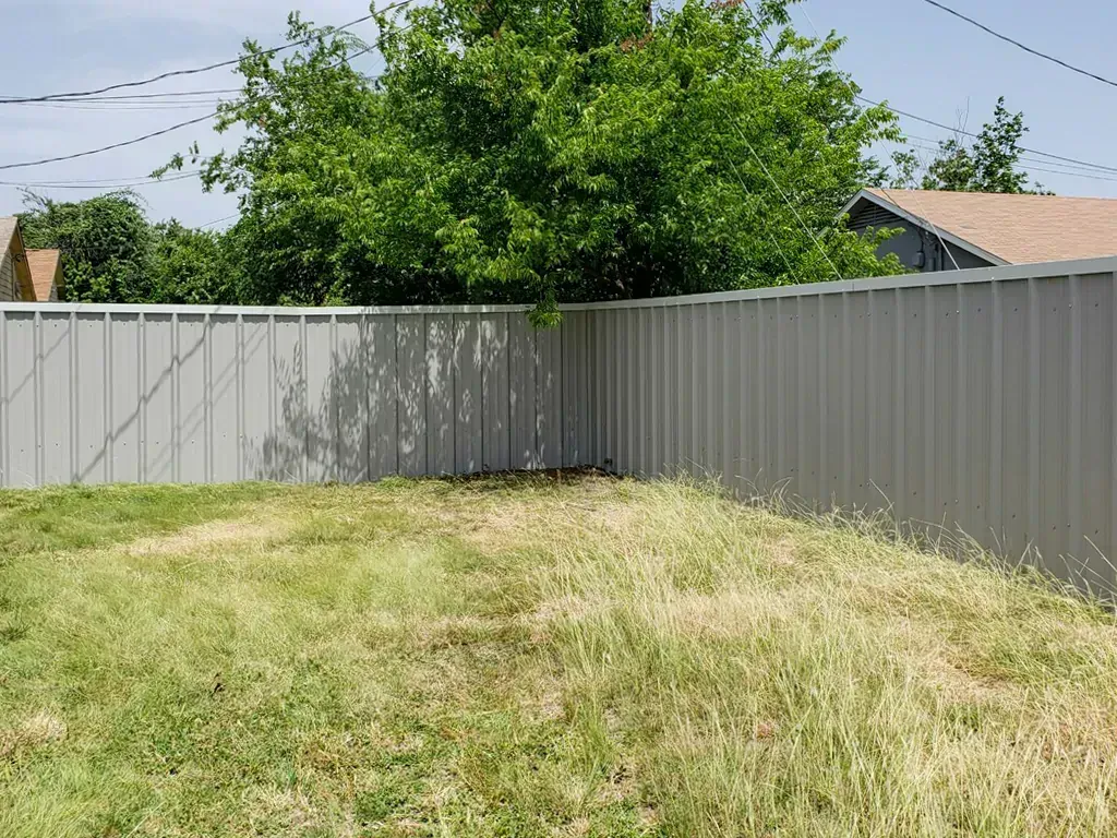 A metal fence encloses a backyard with overgrown grass and a tree, under a blue sky.