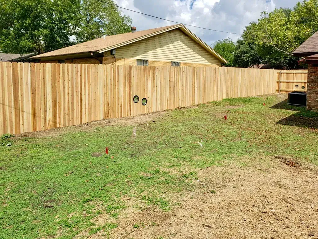 Wooden privacy fence in a backyard; tan boards, green grass, and a beige house visible.