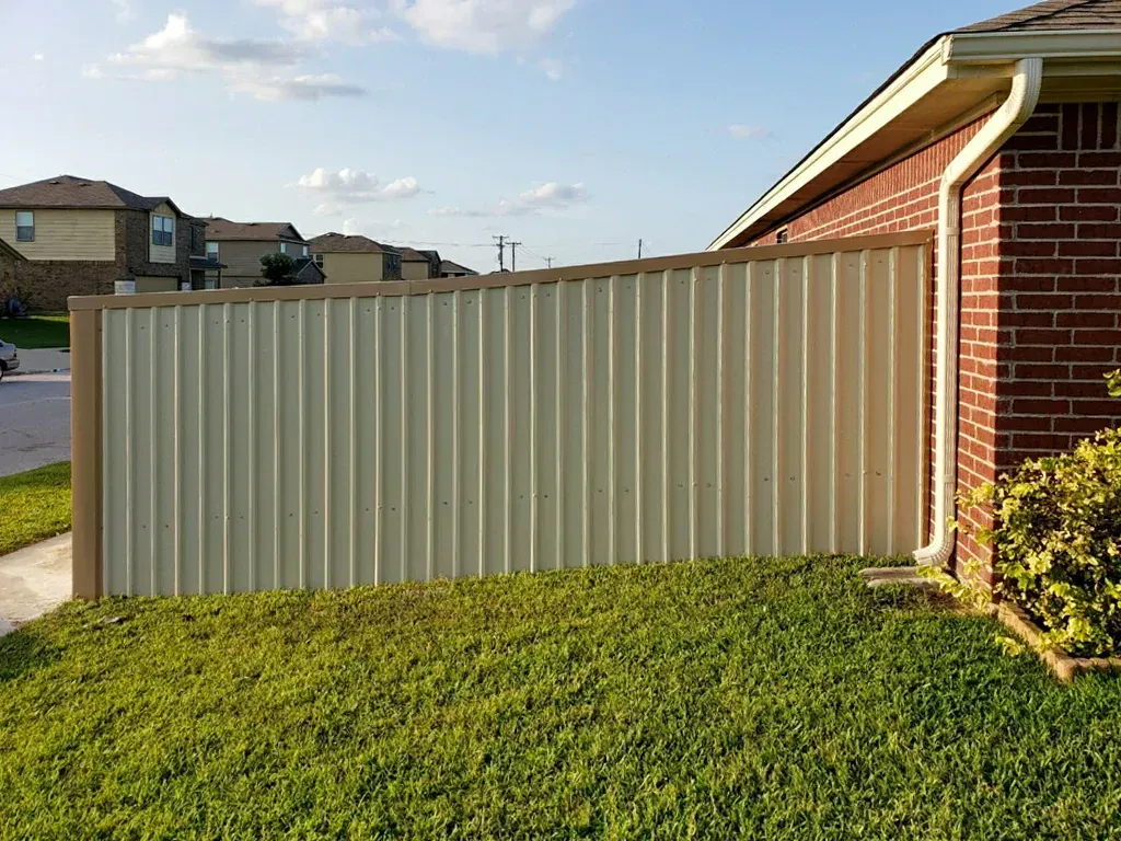 A beige metal privacy fence next to a red brick house in a grassy yard, with neighboring houses visible in the background.