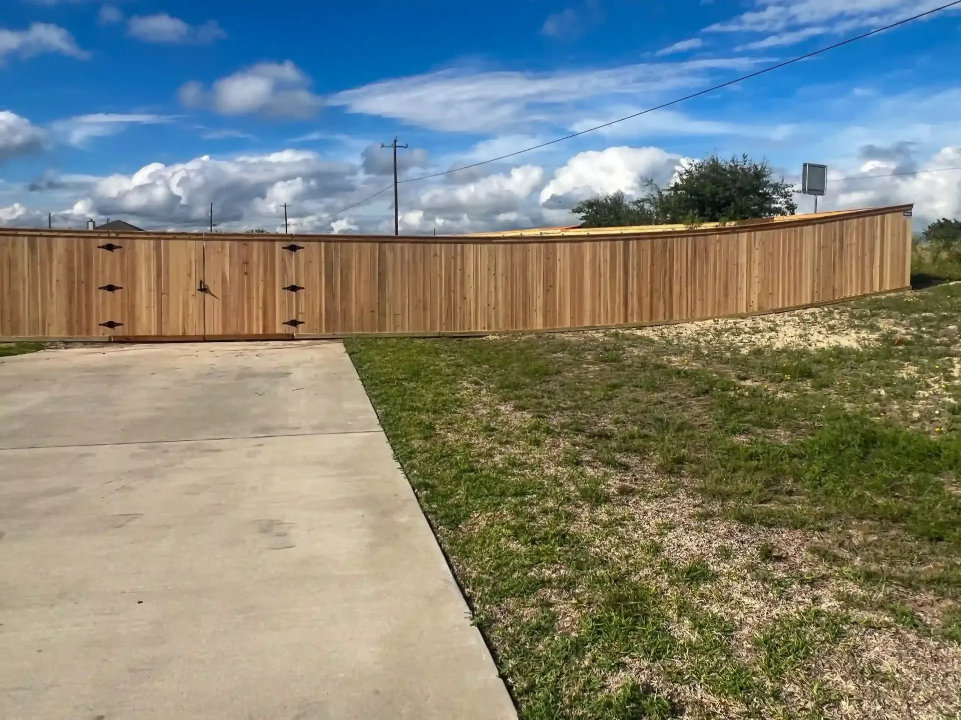 Wooden fence curving along a grassy area under a cloudy sky. A concrete driveway is in the foreground.