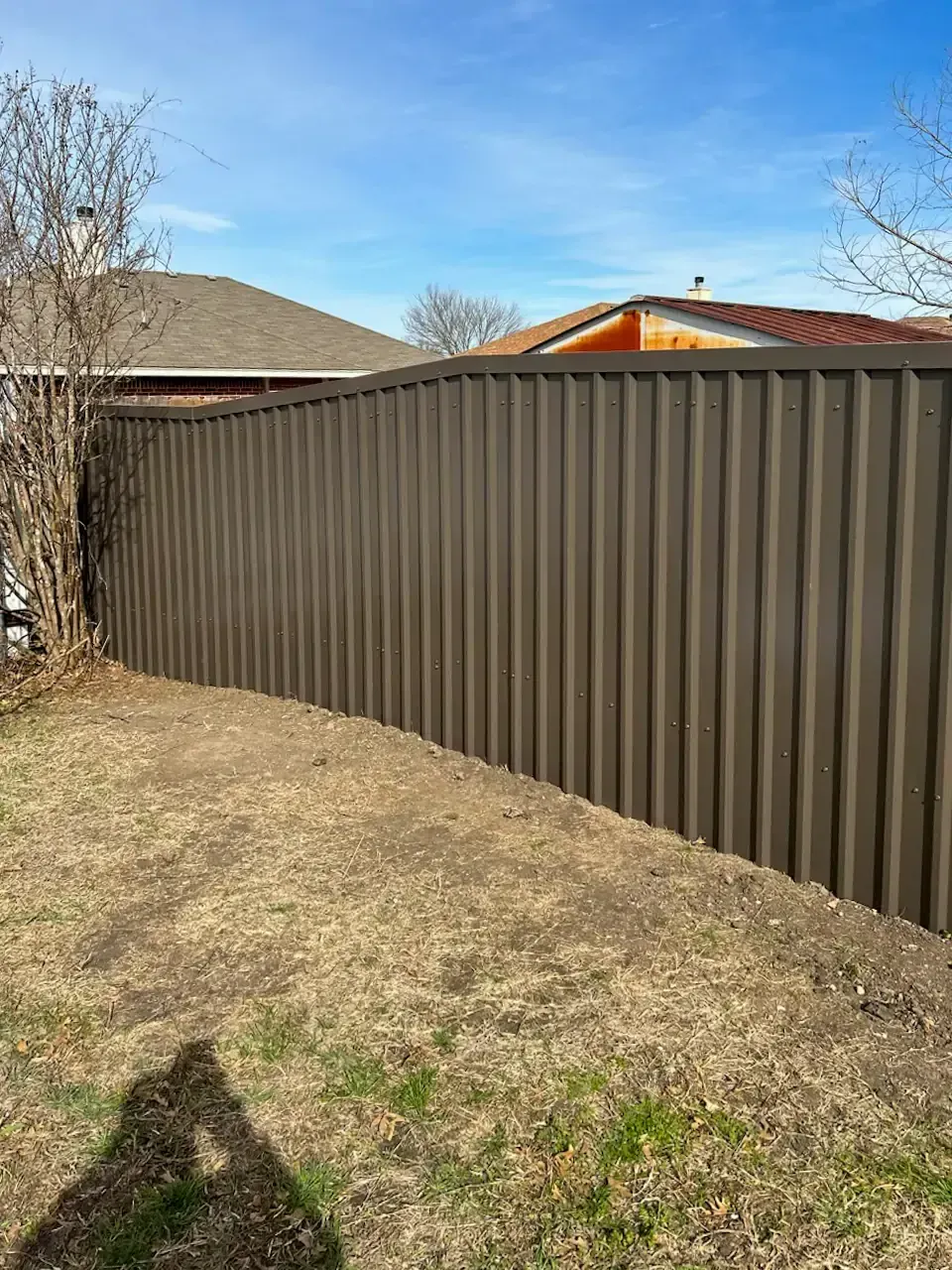 Brown corrugated metal fence in a yard with dry grass and a blue sky.