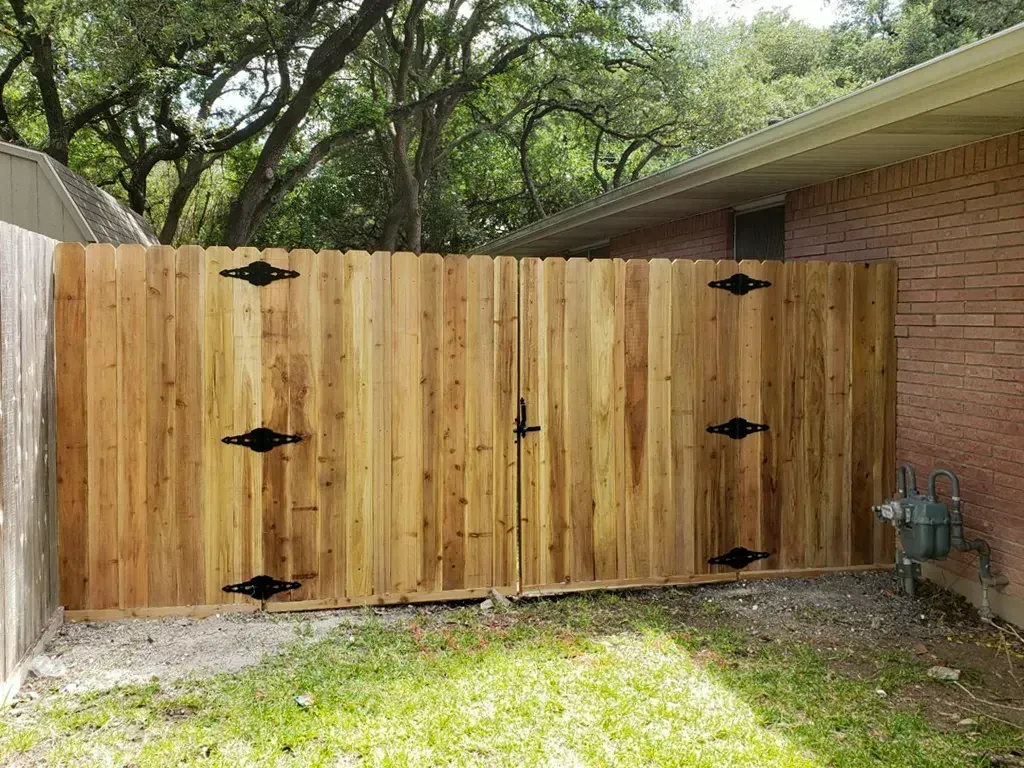 Wooden double gate with black hinges and latch, attached to a fence and a brick house.
