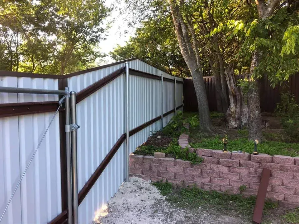 White corrugated metal fence with a brown trim, adjacent to a brick retaining wall and trees.