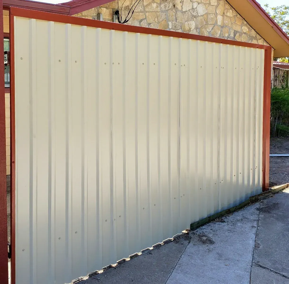 Beige corrugated metal fence with red trim, alongside a stone house and concrete pathway.