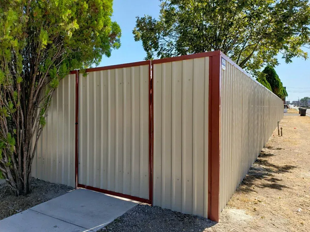 Beige corrugated metal fence with red frame and gate, beside a sidewalk and gravel.