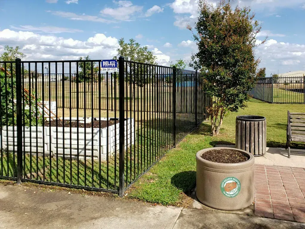 Black fenced-in community garden area with raised beds, a tree, and a concrete planter on a sunny day.