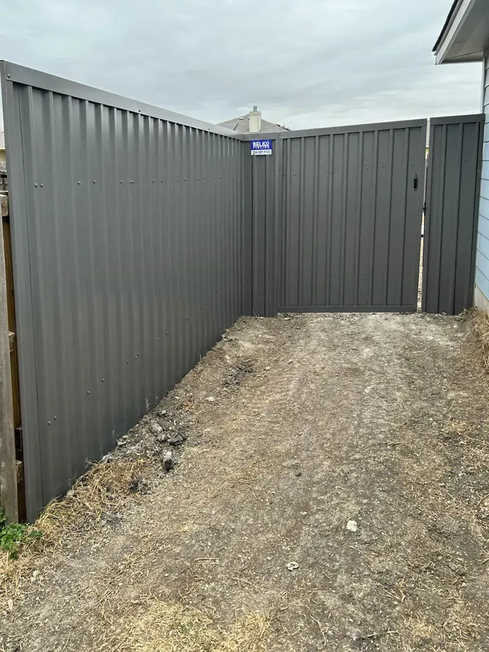 Gray metal privacy fence with gate, enclosing a dirt and gravel area next to a building.