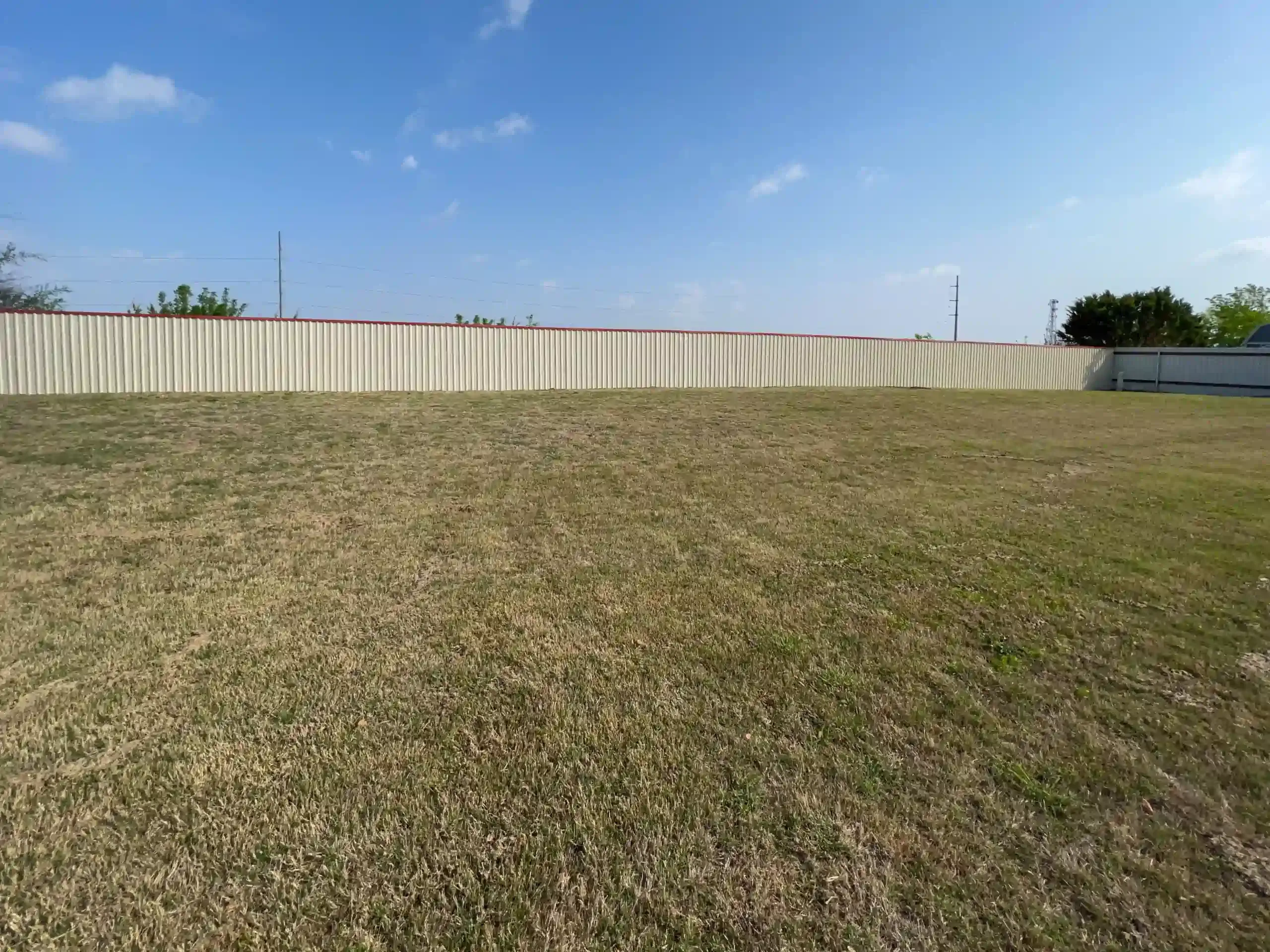 A large grassy field in front of a white wooden fence under a blue sky.