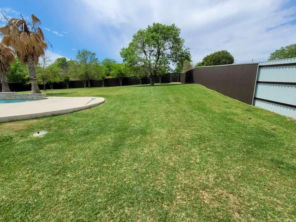 Green lawn with trees and a pool area, enclosed by brown and white fences under a cloudy sky.