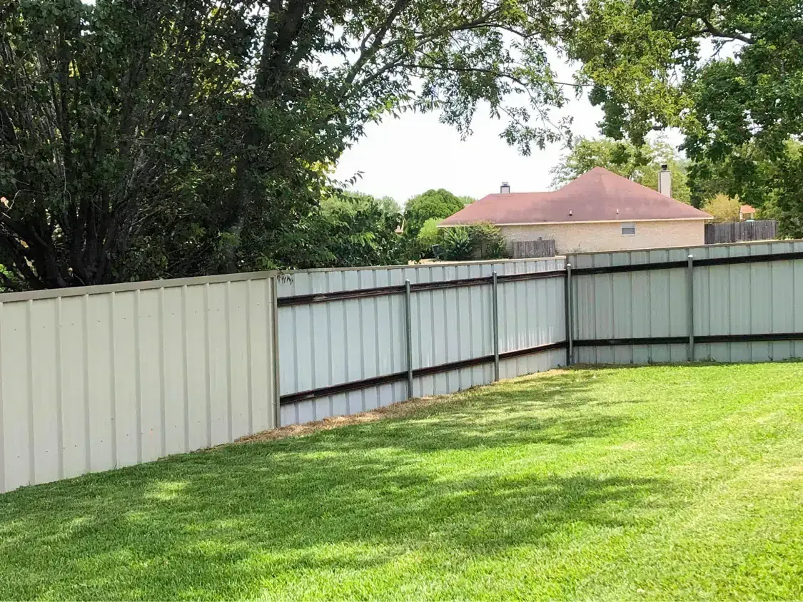 Backyard view with green grass, light-colored fence, and partial view of a house under a sunny sky.