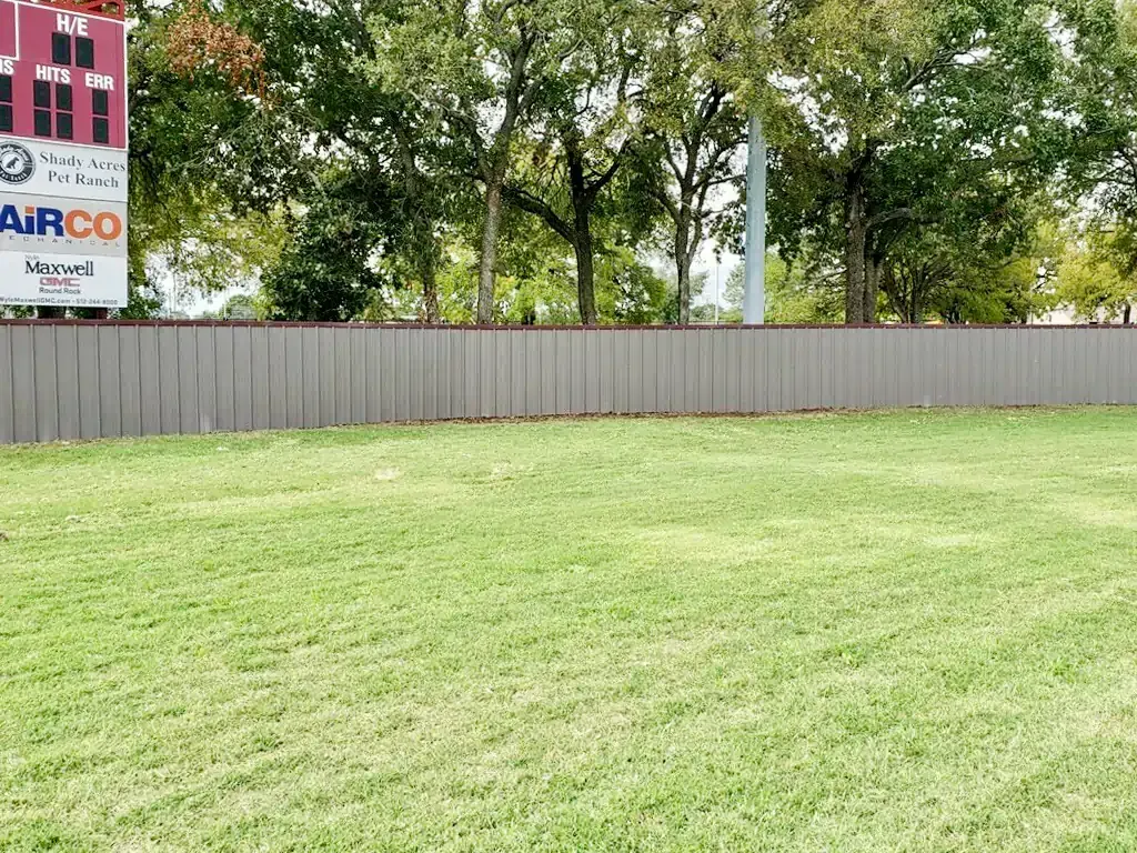 Green grass in front of a gray fence, with trees and a scoreboard in the background.