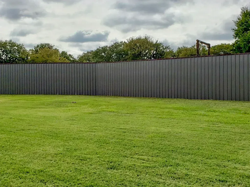 Lush green lawn in front of a tall gray vertical-slat fence, with a cloudy sky and trees in the background.