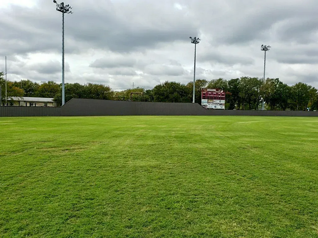 Green sports field with scoreboard, fence, trees, and cloudy sky.