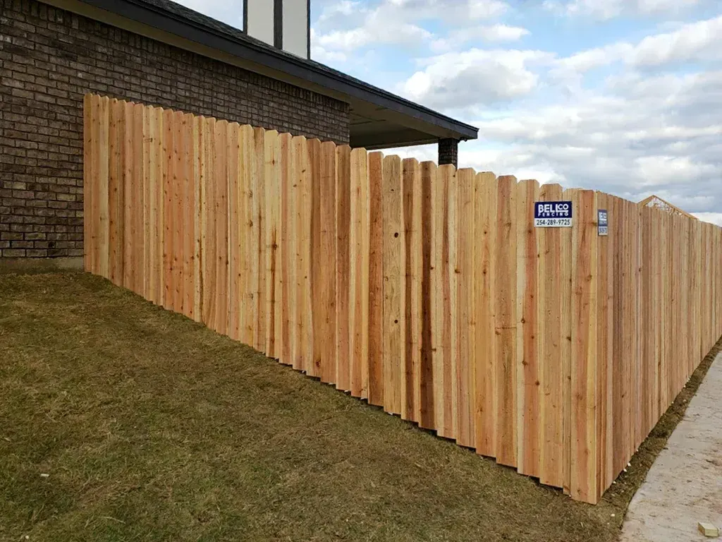 Wooden fence along the side of a building on a grassy slope, with a sidewalk on the right.
