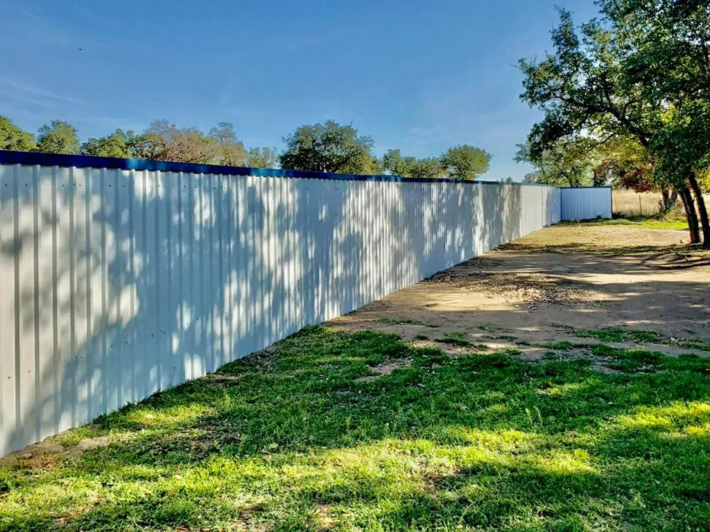 A tall, white metal fence borders a grassy yard, trees in the background under a blue sky.
