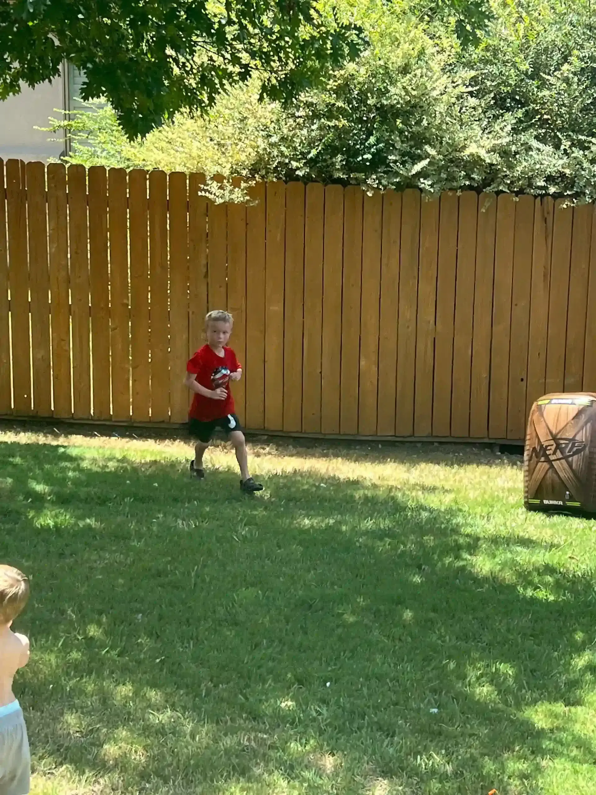 Boy in red shirt running across a grassy yard toward a wooden fence.
