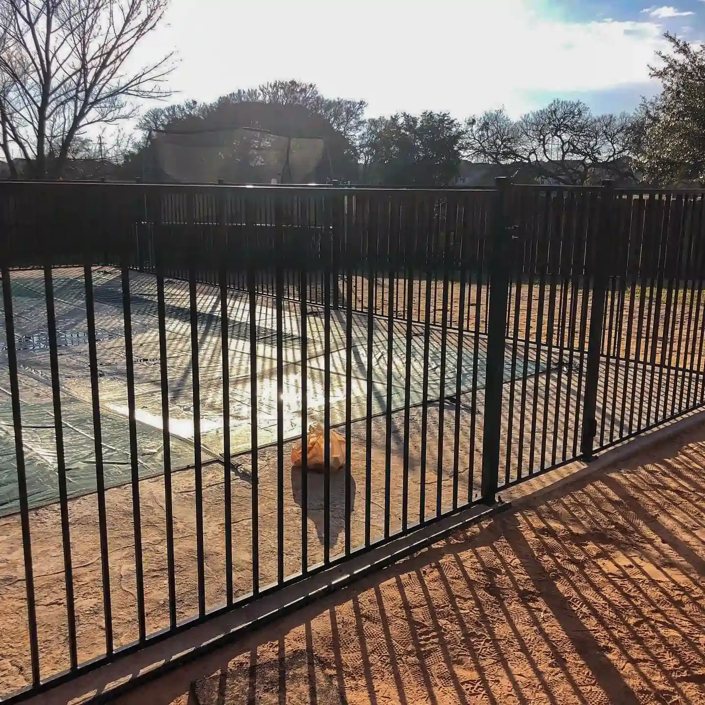 Black metal fence encloses a dirt area with a glimpse of blue, in sunny outdoor setting.
