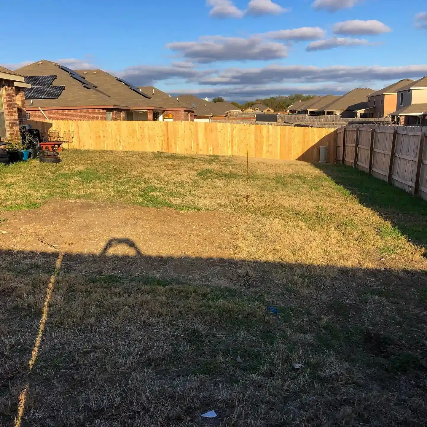 Backyard with a wooden fence, houses, and patchy grass under a partly cloudy sky.
