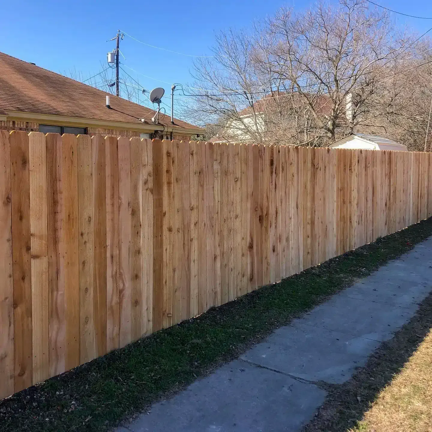 Wooden fence along a paved walkway next to a grassy area, with houses and blue sky in the background.