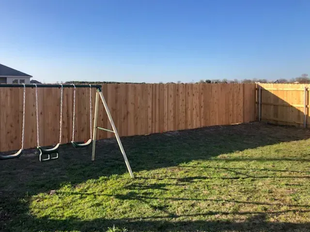 A wooden fence surrounds a grassy backyard with a swing set under a clear blue sky.