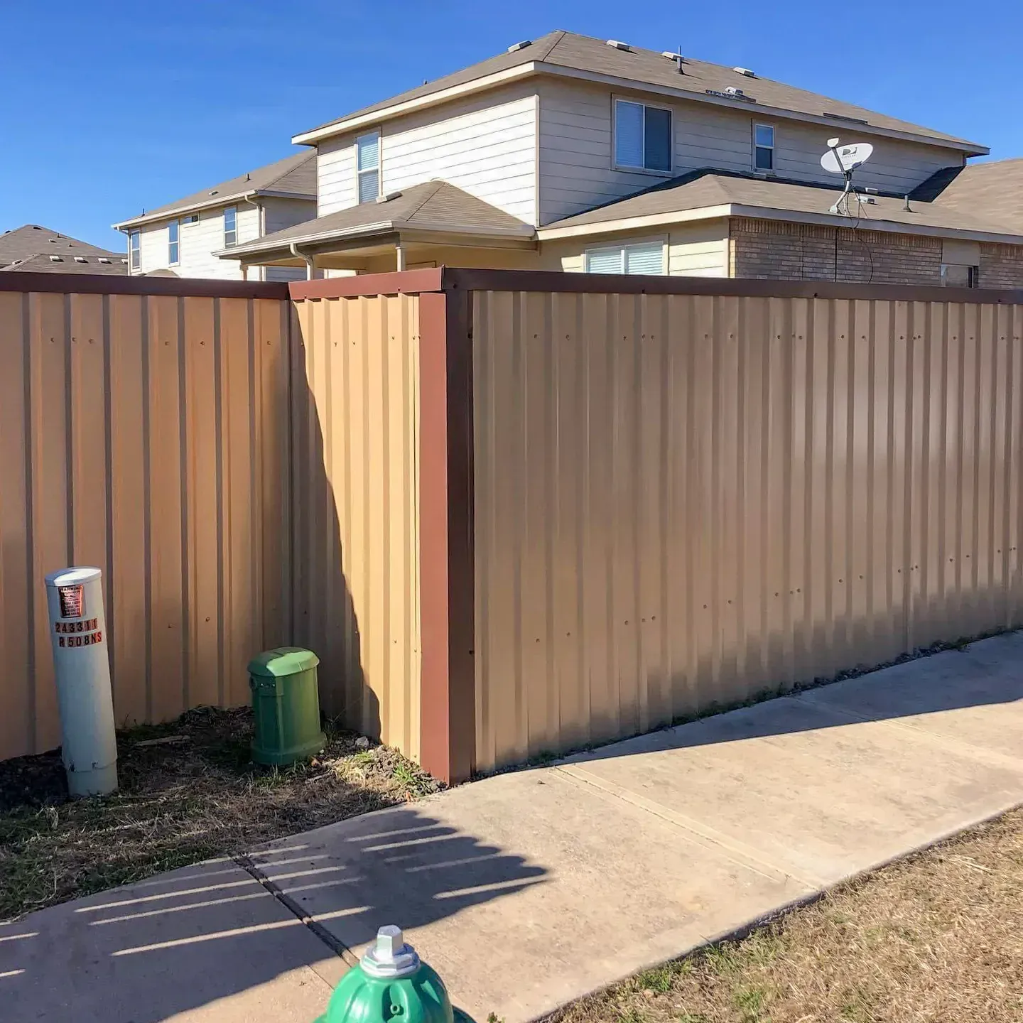 Tan corrugated metal fence with brown trim next to a sidewalk. A two-story house is in the background.