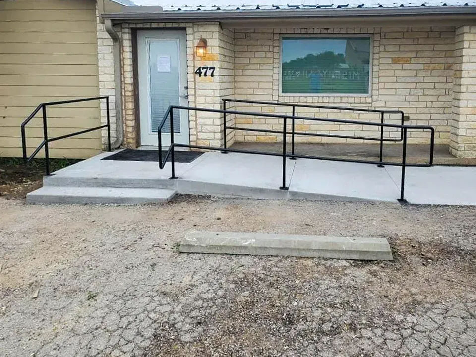 Exterior of a building with a wheelchair ramp and steps leading to the entrance door, gravel ground.