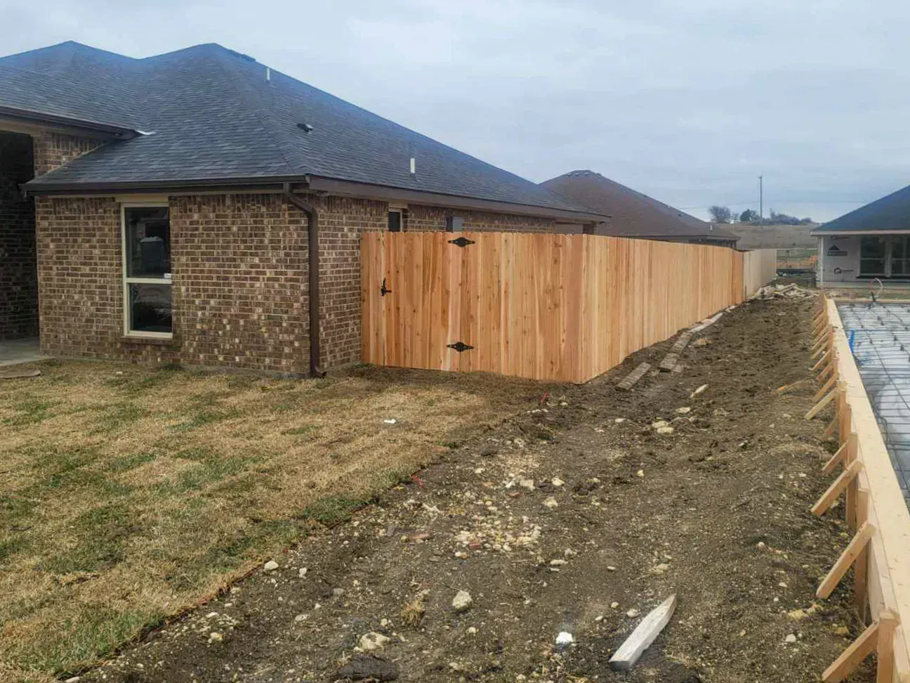 Wooden fence installed alongside a brick house and construction site with dirt and concrete.