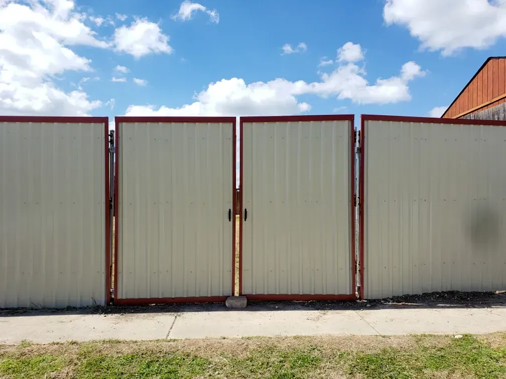 A closed, beige metal gate and fence with brown trim against a partly cloudy sky.