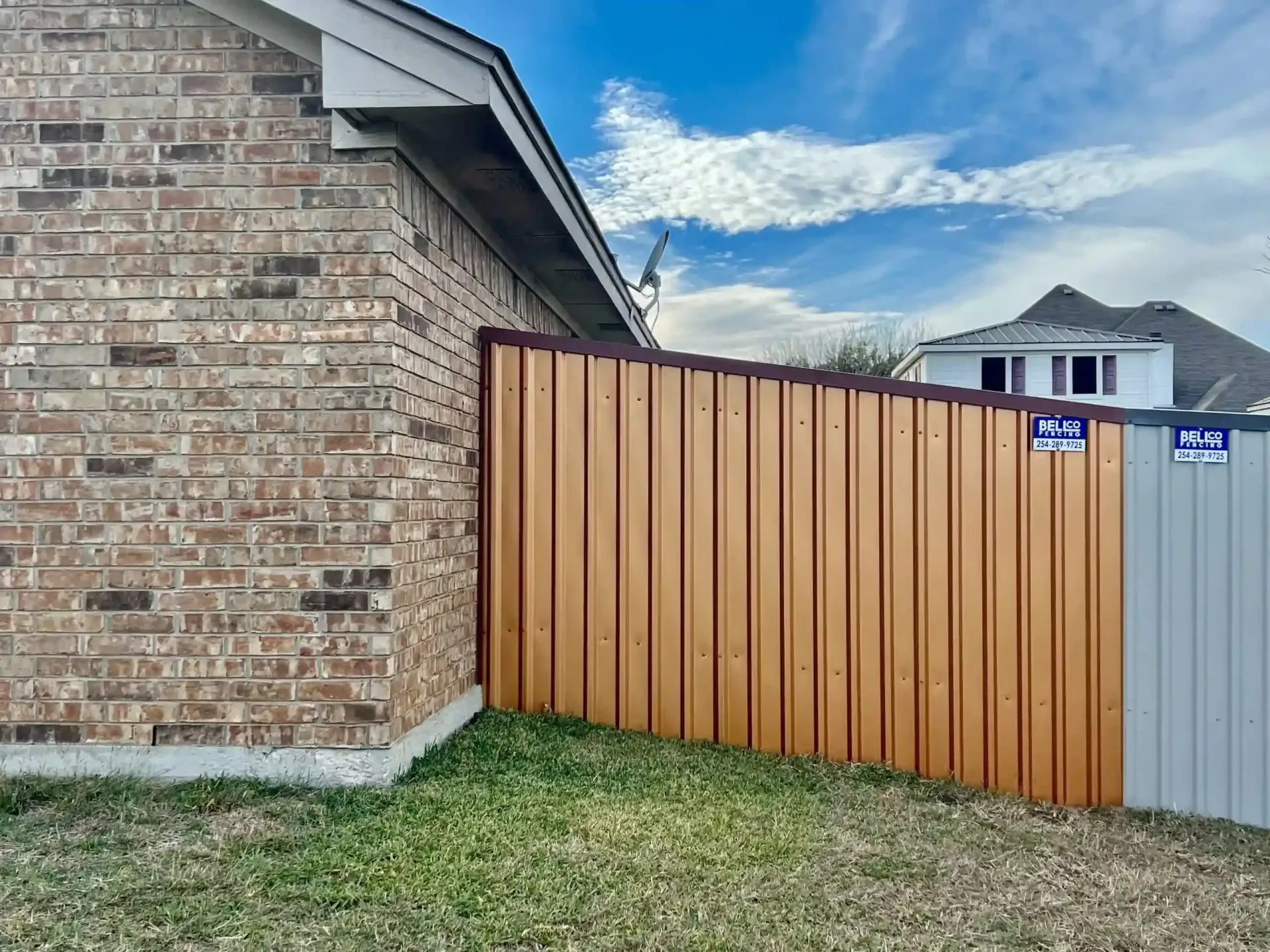 Brown metal fence next to a brick building on a grassy yard under a cloudy sky.
