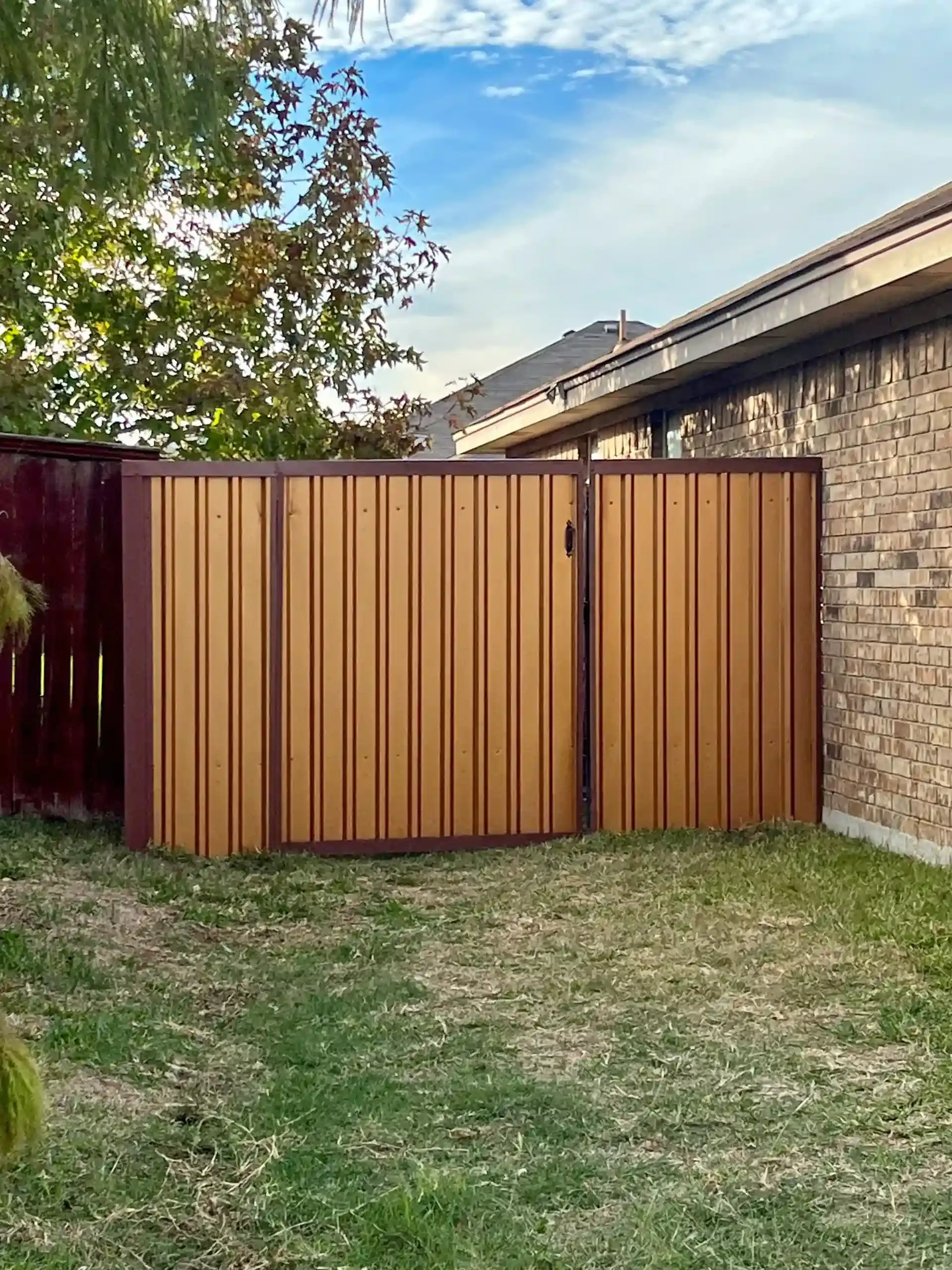 Brown metal fence and gate next to a brick wall in a grassy backyard, under a partly cloudy sky.
