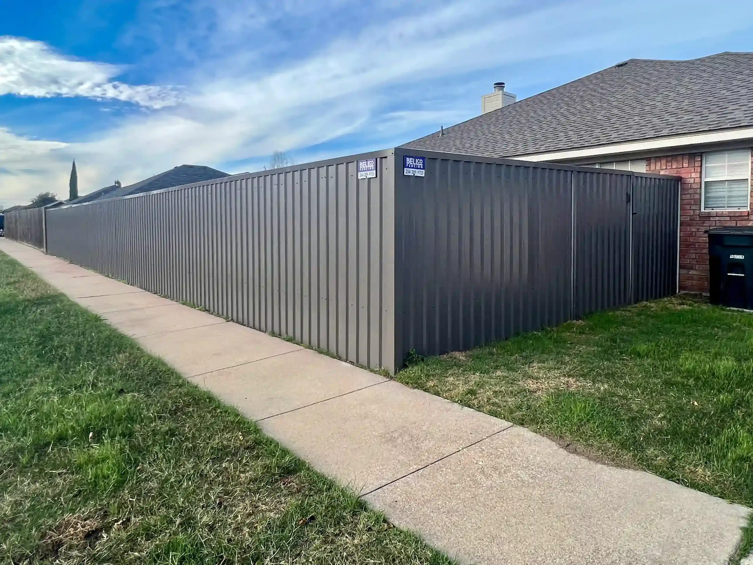 Gray metal fence along a sidewalk next to a house with a brick facade and a gray roof, under a blue sky.