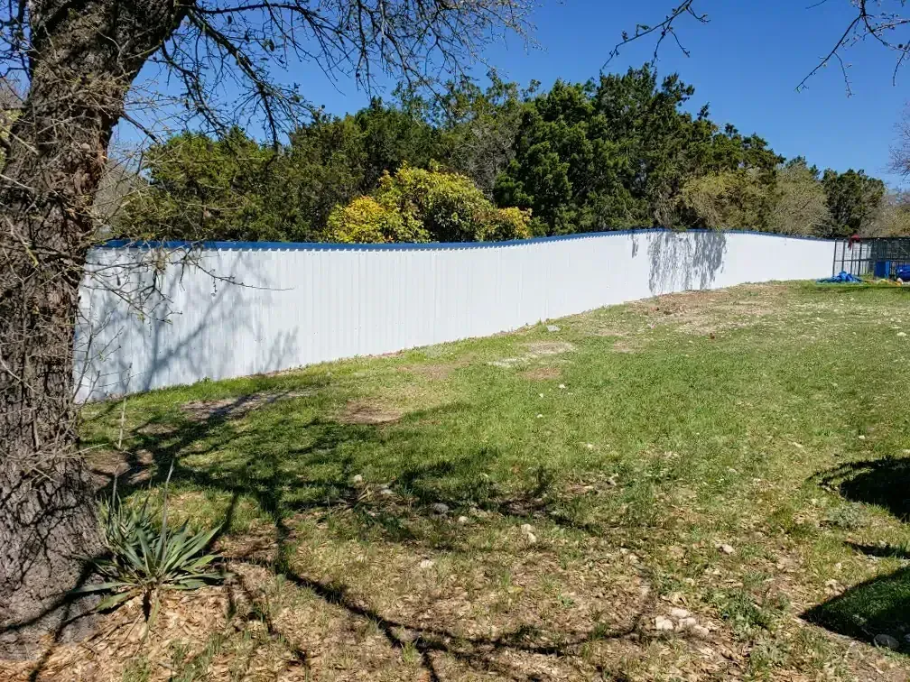 White corrugated metal fence along a grassy yard with trees in the background.