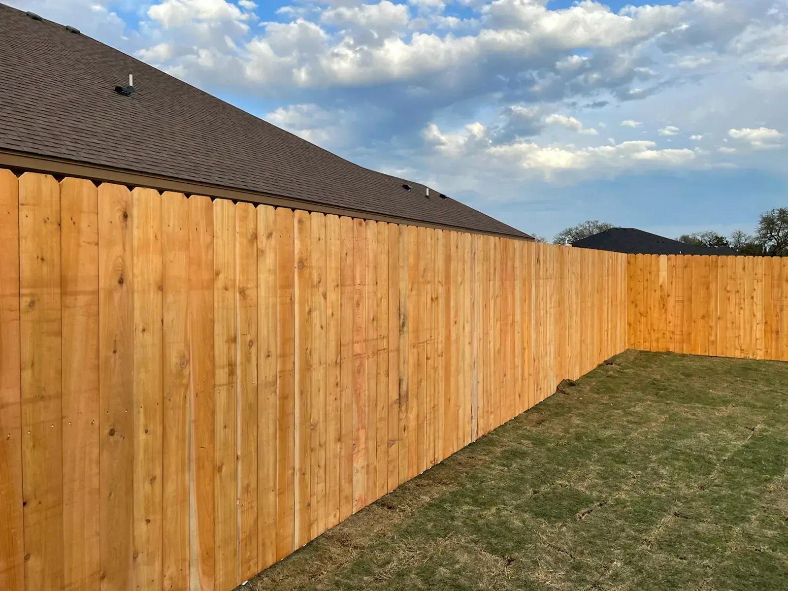 Wooden privacy fence in a backyard with a brown roof and cloudy sky.