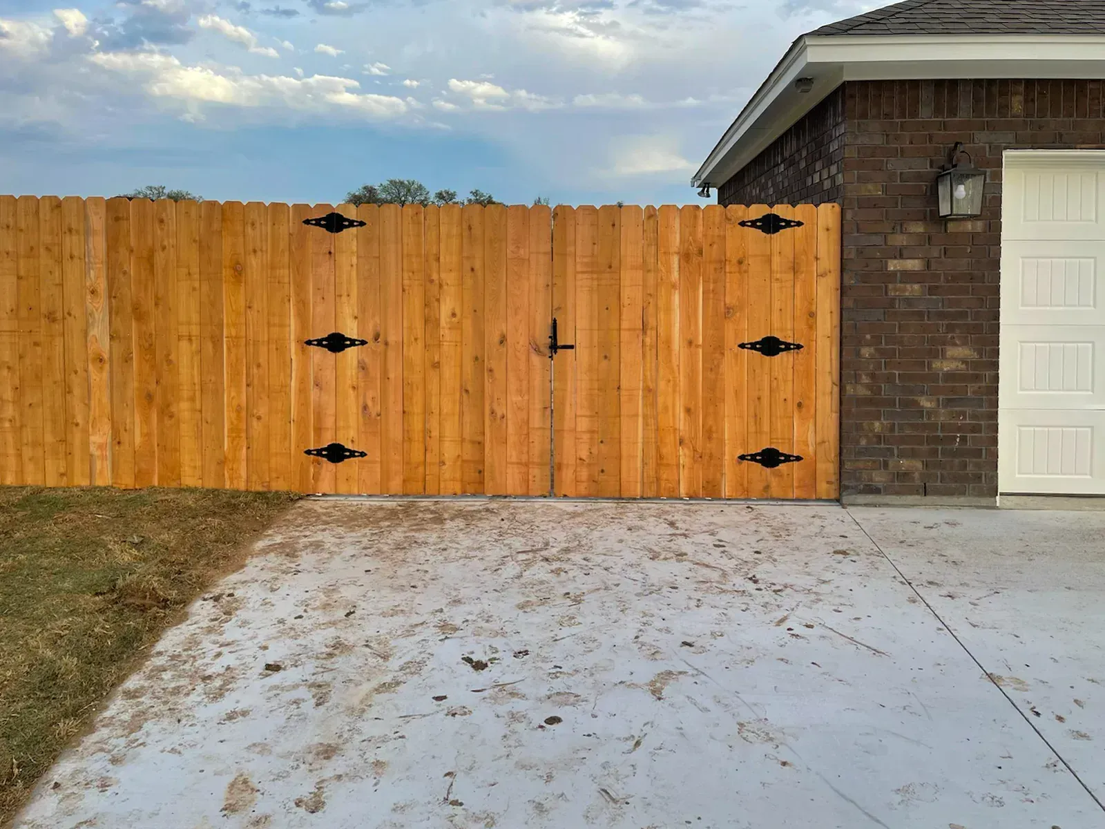 Wooden gate in front of a brick building. Black metal hardware and a lock are visible. Concrete driveway.