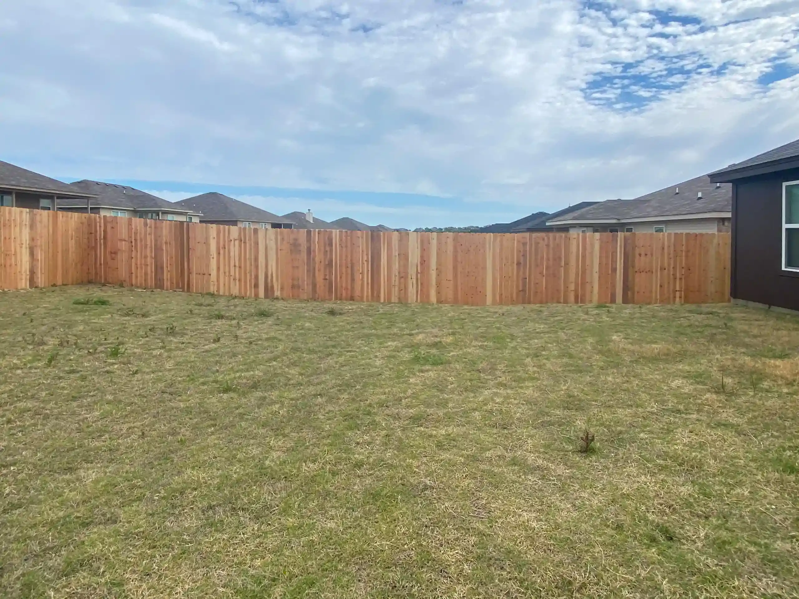 A backyard with a wooden fence. Brown fence surrounds a grassy lawn under a cloudy sky. Houses are in the background.