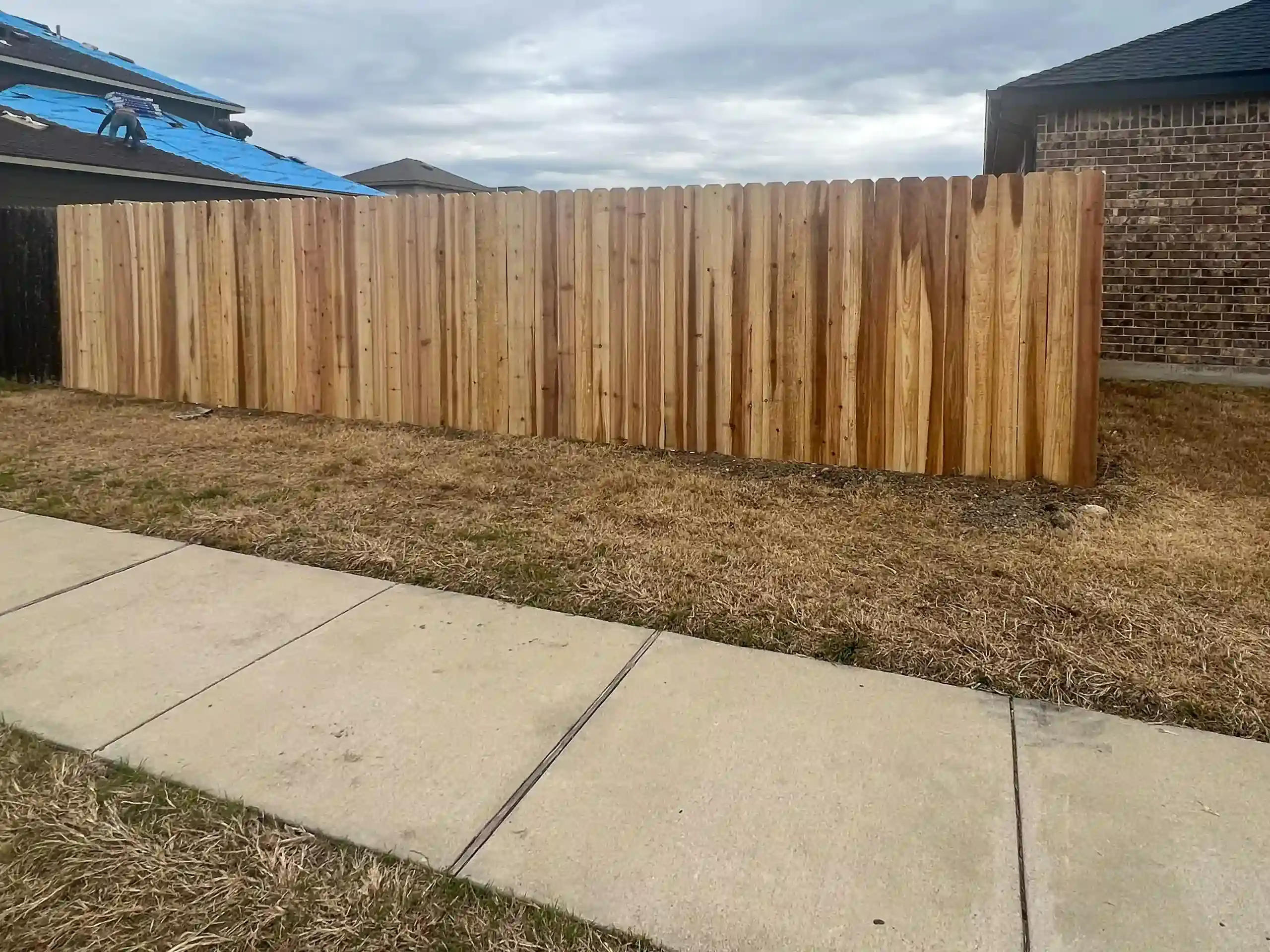 Wooden fence along a grassy yard, beside a sidewalk; cloudy sky in background.