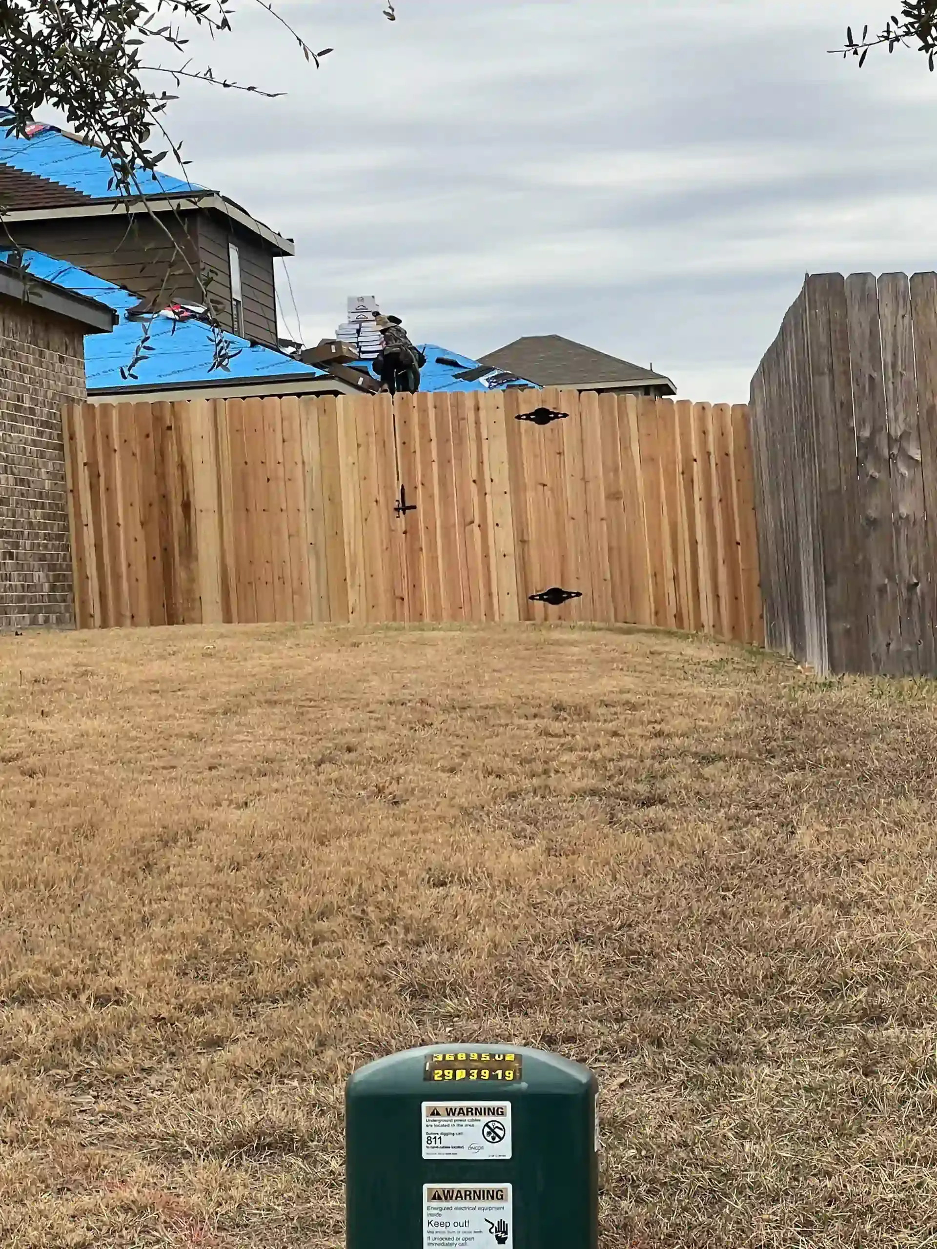 A backyard with a wooden fence, brown grass, and cloudy sky. A green utility box is in the foreground.