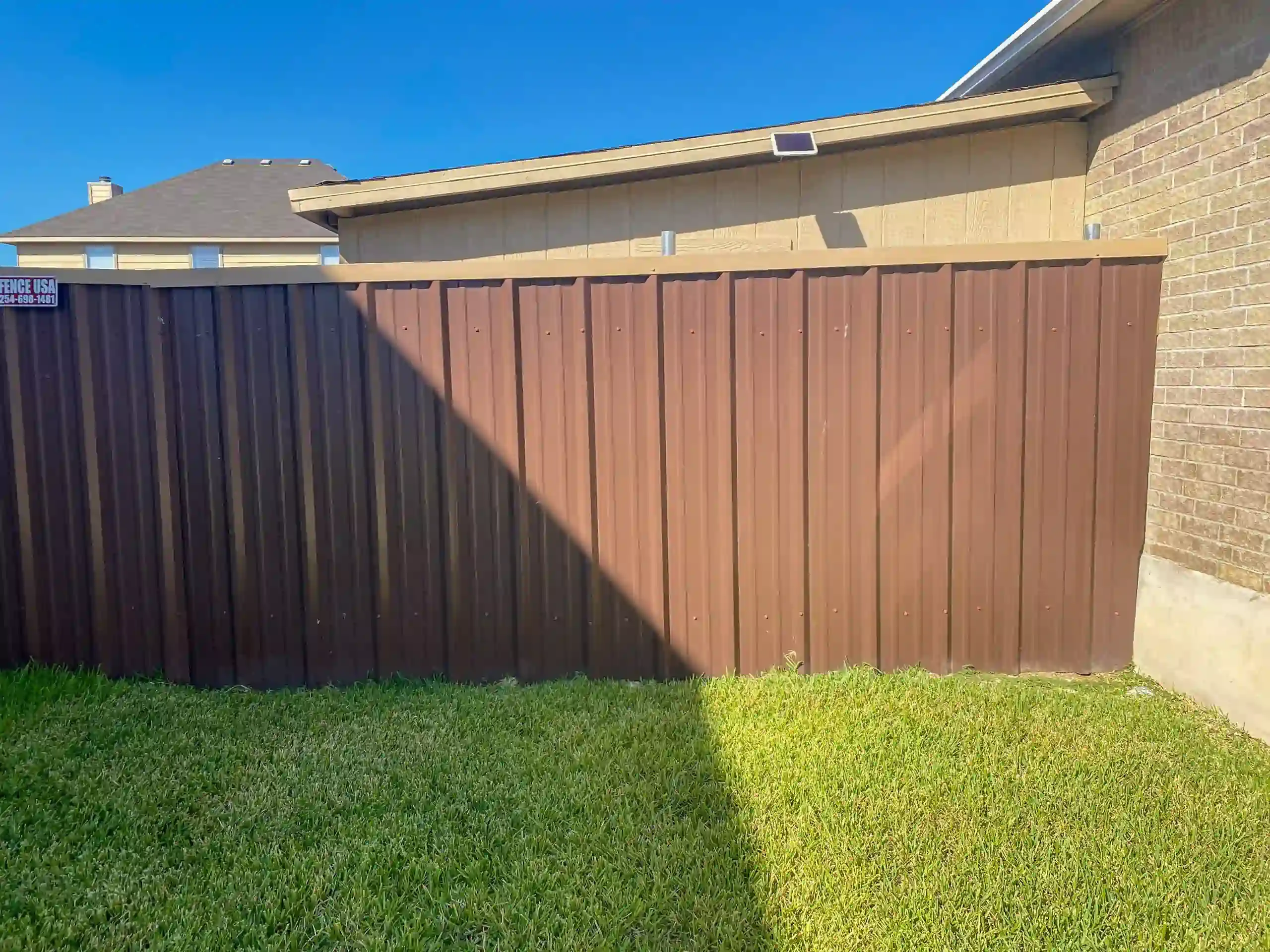 Brown metal fence with green and yellow grass, against a tan wall and roof.