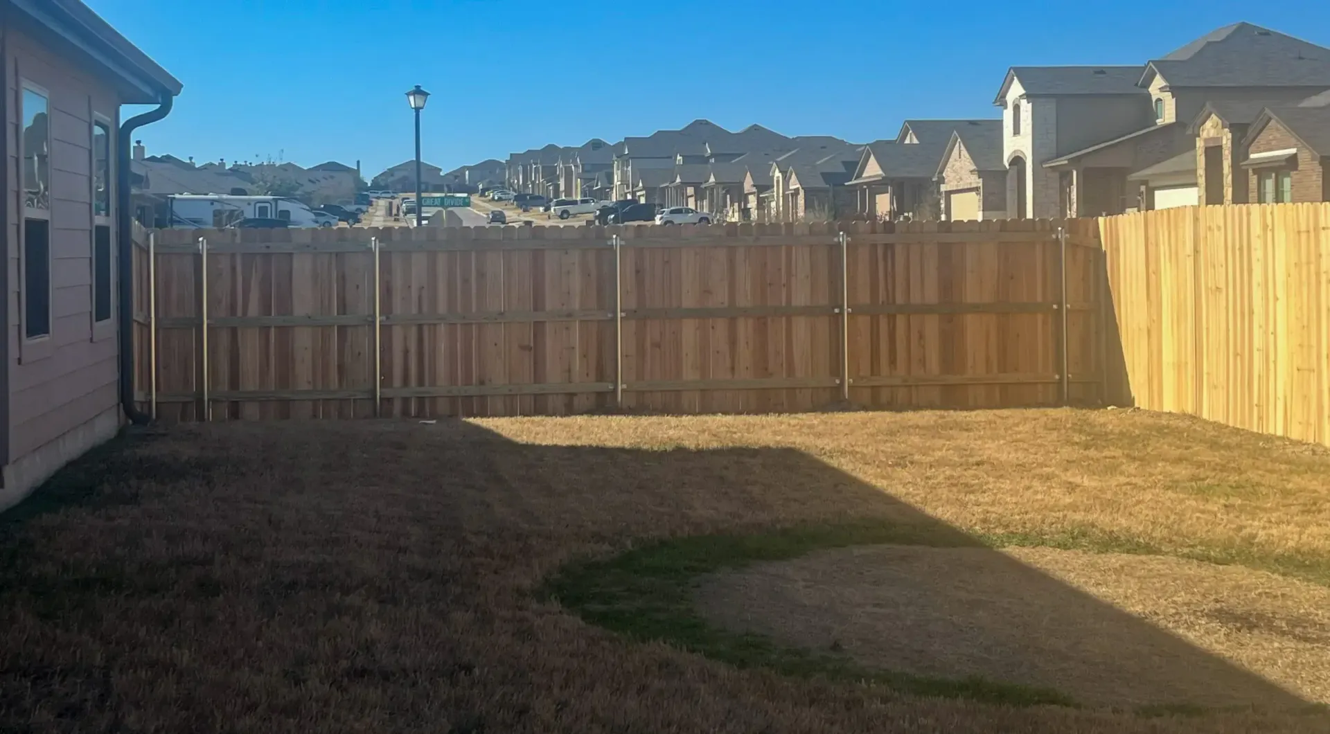 Backyard with brown grass, wooden fence, houses in the distance, and blue sky.