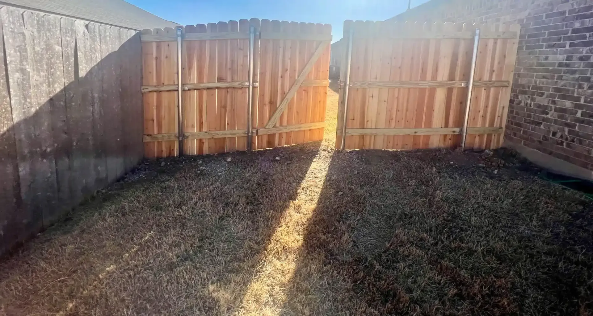 Open wooden gate in a backyard, with sunlight shining through. Brown fence, dark grass.