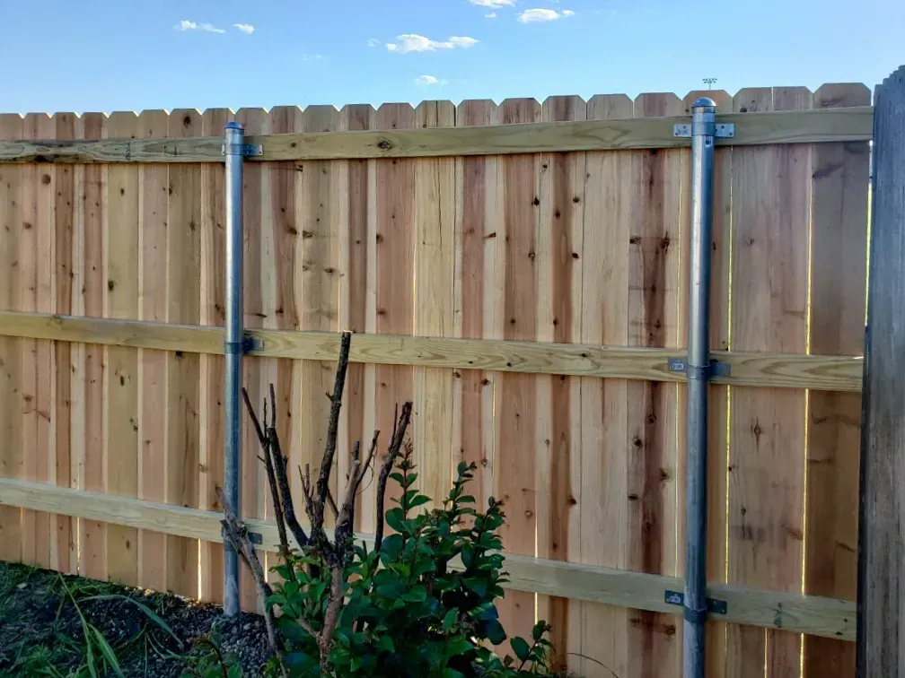 Wooden fence with metal poles and brackets in front of green shrubs under a blue sky.