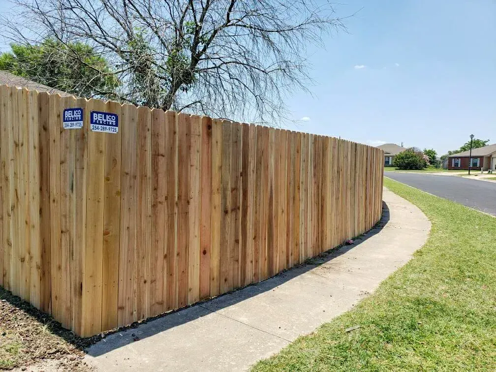 Wooden fence curving along a sidewalk beside a road, blue sky overhead.