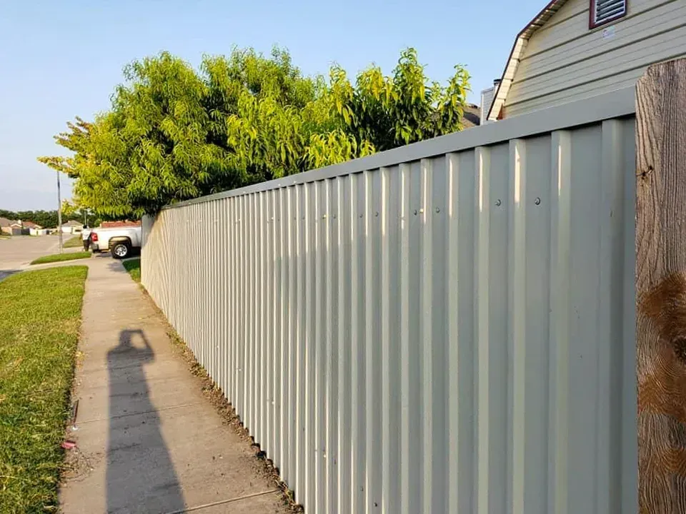 Gray corrugated metal fence along a sidewalk, with a tree and partial house in the background.