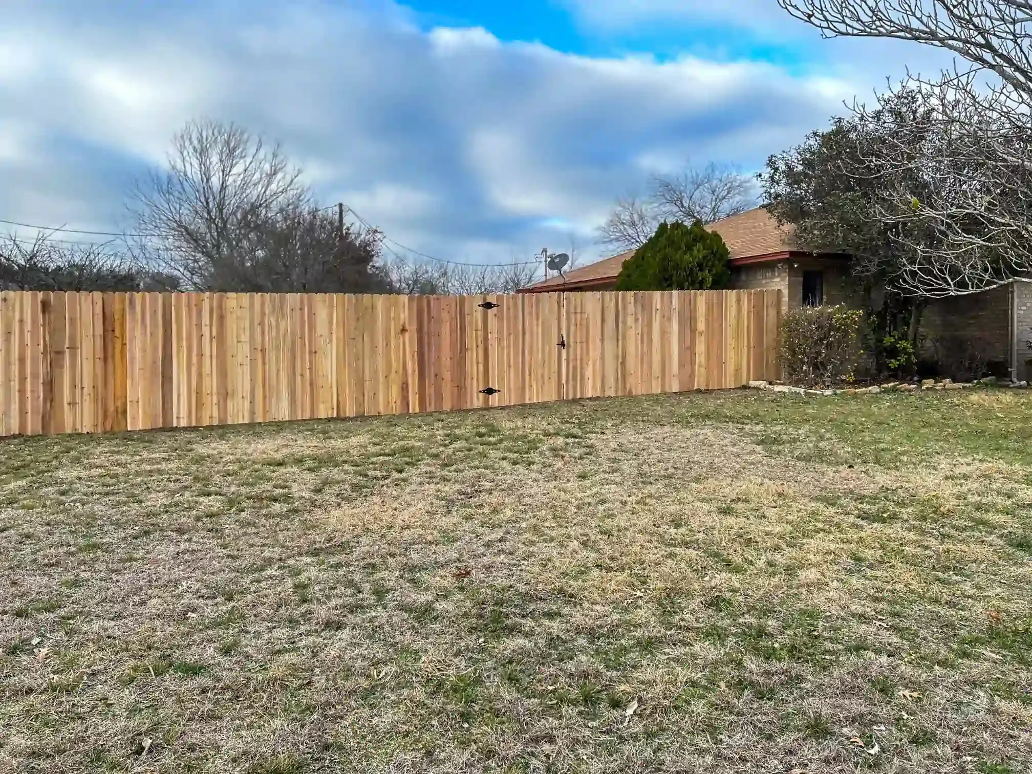 Wooden fence with gate in backyard. Brown grass, cloudy sky, house in the background.