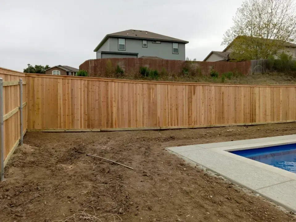 Wooden fence bordering a backyard with a pool. Houses are visible in the background under an overcast sky.