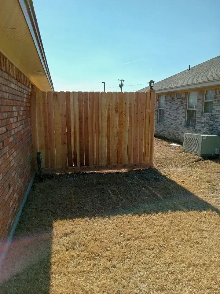 Wooden fence alongside a brick building, enclosing a small yard with brown grass and a neighboring building.